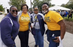 Four people smile and pose together outdoors, wearing shirts in school colors, with a festive crowd and a large tent visible in the background.