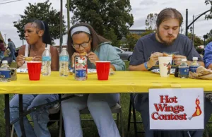 Three participants eat hot wings at a contest, seated at a yellow table. Each has drinks and one displays a cellphone. A sign reads "Hot Wings Contest." Trees and vehicles are visible in the background.