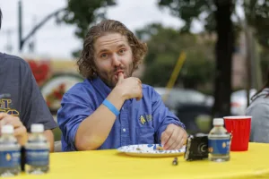 A man with curly hair playfully licks his finger while sitting at a table with a yellow tablecloth, surrounded by water bottles and a red cup. He is wearing a blue shirt with a logo.