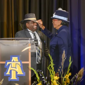 A joyful interaction between two individuals at a podium, with one person adjusting the other's hat. They are surrounded by flowers and a backdrop featuring a logo. Both wear formal attire and appear to be celebrating an occasion.
