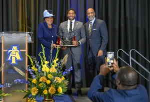A woman in a blue suit and hat presents a man in a gray suit with an award. Another man in a blue suit smiles, while an attendee captures the moment on a phone. A colorful floral arrangement decorates the stage.
