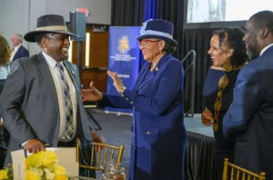 A group of four people in formal attire engage in a joyful conversation at an event. Two women and two men smile and laugh, with a banner visible in the background. A table with floral arrangements is in the foreground.