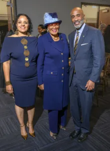 Three individuals pose together, dressed in stylish blue outfits. The woman on the left wears a navy dress with a statement necklace, while the central figure, an older woman, sports a chic blue coat and hat. The man on the right is dressed in a tailored suit.