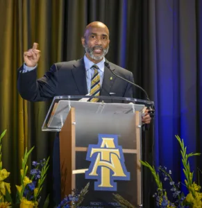 A man with a beard speaks at a podium decorated with flowers. He is wearing a suit and tie, gesturing with his right hand, while addressing an audience. The backdrop features dark curtains.
