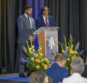 Two young men stand at a podium with flowers in the foreground. One is speaking while the other looks on attentively. The backdrop features a black curtain and the logo of North Carolina A&T State University.