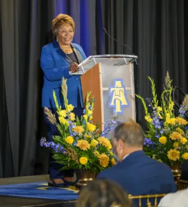 A woman in a blue suit speaks at a podium adorned with flowers. She smiles and gestures, while audience members are seated in front of her, engaged in the event.