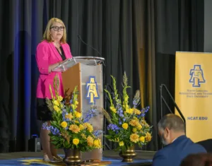 A woman in a pink blazer speaks at a podium adorned with flowers, representing North Carolina Agricultural and Technical State University. Audience members are visible in the foreground.