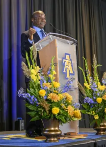 A speaker passionately addresses an audience from a podium adorned with flowers. Vibrant yellow and blue blooms flank the podium, with a logo of a university visible.