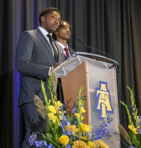 Two young men speaking at a podium with the North Carolina A&T State University logo. The podium is surrounded by colorful flowers. One speaker is wearing a suit, while the other is dressed in a blazer and tie.
