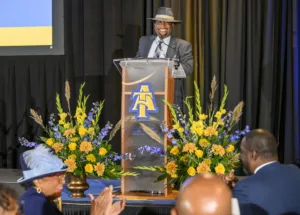 A speaker smiles at a podium adorned with vibrant floral arrangements during an event. Attendees are seated nearby, clapping in appreciation. The backdrop features the logo of North Carolina Agricultural and Technical State University.