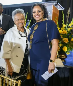 Two women pose together, smiling, at an event. One wears a white jacket over a black dress, the other in a navy blue dress with bold earrings. A floral arrangement is visible in the background, adding color to the setting.