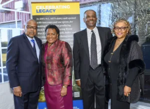 Four people pose together, smiling at an event. They stand in front of a backdrop with text about N.C. A&T's dairy unit and a celebration of legacy.