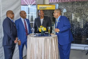 Four men in formal attire converse around a small table decorated with flowers and drinks. The background features a banner celebrating a legacy.