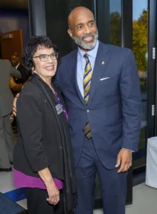 Two smiling individuals pose together in a formal setting. One person wears glasses and a black jacket over a purple top, while the other is in a blue suit with a striped tie.