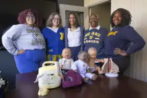 Five women stand together, smiling, in a classroom setting. They are grouped around a table displaying educational models and baby dolls, suggesting a focus on health or education training.