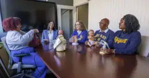 Six women are seated around a table, engaging in discussion. They hold various items including baby dolls and anatomical models, with a relaxed atmosphere in a well-lit room.