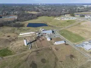Aerial view of a rural landscape featuring several buildings, silos, and a pond, surrounded by fields and wooded areas. The skyline of a city is visible in the background.