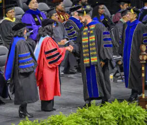 A graduate in a black gown with a colorful stole shakes hands with an official in a red gown at a graduation ceremony. Other graduates and faculty members are seated in the background.