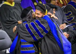 Graduates in academic regalia embrace joyfully, celebrating their achievements at a commencement ceremony. Friends and family are present in the background, sharing in the moment.