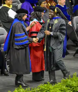 Three individuals in academic regalia celebrate at a graduation ceremony, shaking hands and smiling. A backdrop of seated attendees creates a festive atmosphere.