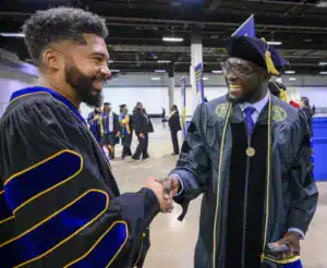 Two men in graduation gowns shake hands and smile at each other in a celebratory setting. Graduates can be seen in the background, and the atmosphere is joyful and festive.