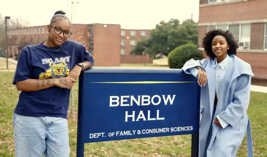 Two women stand beside a sign for Benbow Hall, Department of Family & Consumer Sciences. One wears glasses and a t-shirt, while the other is in a light blue coat, both smiling in front of the building.