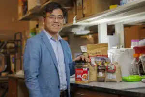 A man smiles while standing beside a collection of food products, including popcorn, grains, and flours, in a laboratory setting. He is dressed in a light blue blazer and has glasses.