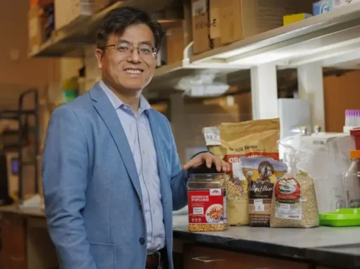 A man smiles while standing beside a collection of food products, including popcorn, grains, and flours, in a laboratory setting. He is dressed in a light blue blazer and has glasses.