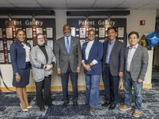 A group of six individuals stands in front of a wall displaying framed patents, celebrating a distinguished achievement in a professional setting. Colorful balloons are also present.
