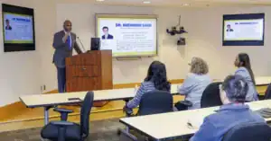 A speaker presents information to an audience in a classroom setting, with slides displayed on a screen featuring details about Dr. Shengmin Sang and his professional background.