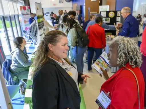 Two women engage in conversation at a bustling event. One wears a black blazer and gestures while the other holds a brochure, surrounded by people and informational displays.