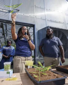 A woman holds a plant with roots while speaking into a microphone. Two people stand nearby, with one holding a jar of seedlings. In the background, greenhouse equipment is visible.