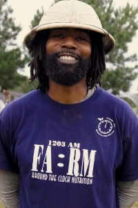A smiling man with a beard and dreadlocks wears a blue t-shirt and a wide-brimmed hat. He stands outdoors in front of trees, exuding a friendly demeanor.