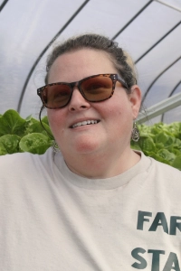 A woman wearing sunglasses smiles in a greenhouse filled with vibrant green lettuce. She has medium-length hair and is dressed in a light-colored shirt with "FARM" printed on it.