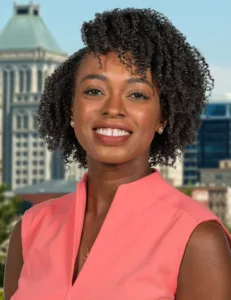 Smiling woman with curly hair wears a coral top. City skyline in the background.