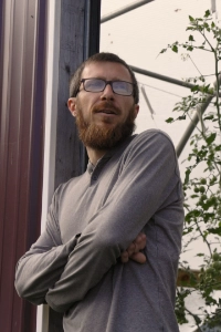 A bearded man with glasses stands with his arms crossed, looking thoughtfully to the side. He's wearing a long-sleeved gray shirt, and there are plants and structures in the background.