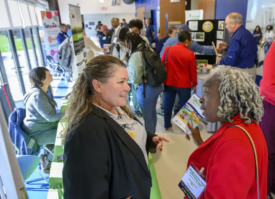 Two women engage in conversation at a bustling event. One wears a black blazer and gestures while the other holds a brochure, surrounded by people and informational displays.
