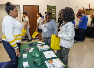 People engaging at a table with informational materials and giveaways. Two women are discussing while holding yellow bags. Others are seen in the background exploring the event.