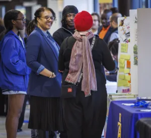 A group of people at an indoor event, engaging in conversation. One woman in a blue blazer smiles as she talks to a person wearing a red hat. Others are also part of the discussion, with informational displays visible.