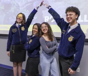Four young adults are posing and smiling. They are wearing blue jackets with emblems, with two forming an arch with their arms. The background features a presentation screen.