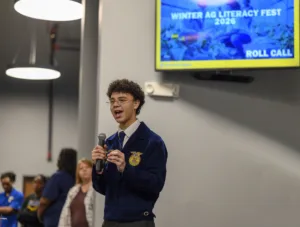 A young man with curly hair and glasses speaks into a microphone, wearing a blue jacket with an insignia. In the background, other attendees listen, and a screen displays "WINTER AG LITERACY FEST 2026 ROLL CALL.