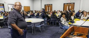 A speaker addresses a large audience seated at tables in a conference room. Attendees appear engaged, with some looking up and others focused on their notes and materials.