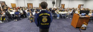 A diverse group of students listens attentively to a speaker wearing a blue jacket with "North Carolina Association" on the back. Tables are set up for discussion in a spacious venue.