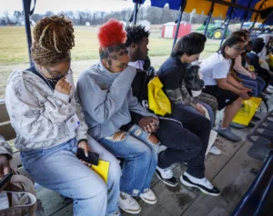 A group of young people sitting closely on a bench, some looking pensive and covering their mouths or faces, with a blurred outdoor scene in the background. Most hold yellow bags.