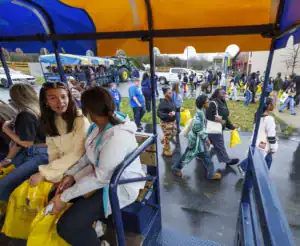 A group of students are either boarding or disembarking from a wagon. Some are walking with yellow bags, while others are seated and chatting inside the wagon. The setting appears to be a school or event location on a rainy day.