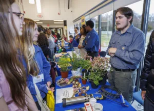 Two young women engage with a presenter at an event. The table features various plants and tools, while other attendees are visible in the background, participating in discussions or browsing the displays.