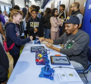 A girl plays rock-paper-scissors with a man at a table featuring robotic arms and tablets. Students and adults engage in discussions in the background during a lively event.