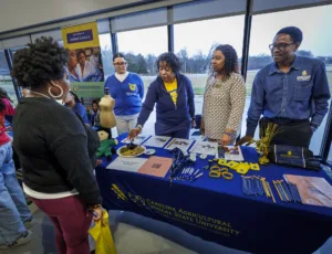 A group at a university booth engages with visitors. Tables display promotional materials, pens, and other items. There’s a mannequin and a doll dressed in green clothing. Several people are interacting, sharing information about animal sciences.