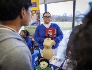 A person with glasses and a blue sweater holds a book entitled "A Career That Makes an Amazing Difference" while speaking to a group. Educational materials and a mannequin are visible on the table.