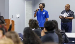 A speaker in a blue shirt holds a microphone, engaging with an audience. Another man in glasses stands nearby, holding a booklet. Attendees are seated, some raising hands.